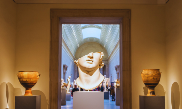 View of a museum gallery centered on a damaged marble bust, missing everything above the nose.