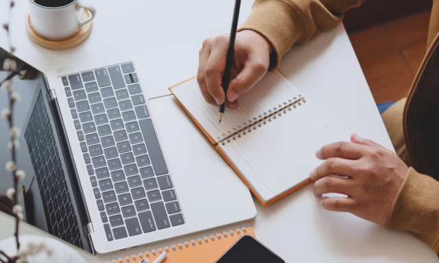 Overhead view of a person writing in a notebook next to a laptop.