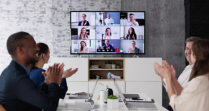 October 29 – 30 | AAM Future of Museums Summit 2024 Four co-workers sitting at a worktable, applauding a large monitor with a webinar in process.