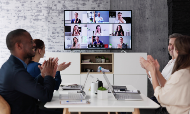 Four co-workers sitting at a worktable, applauding a large monitor with a webinar in process.