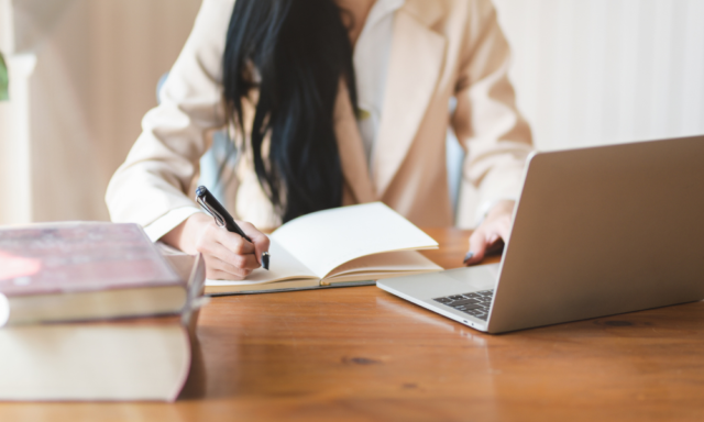 Torso view of a woman writing in a notebook while working at a laptop