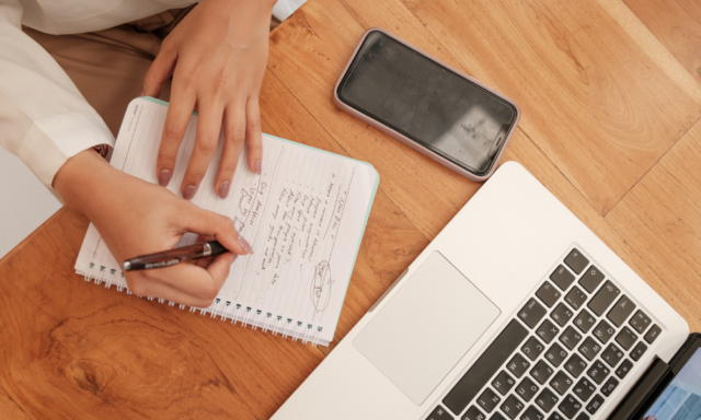 View from above of a personal writing in a notebook with a cell phone and laptop nearby.