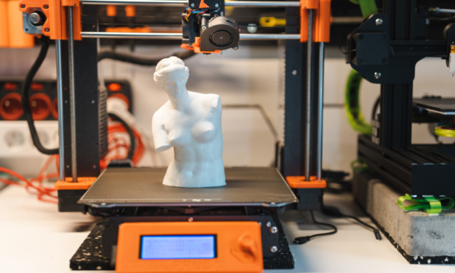 an orange and black 3d printer printing a replica of a classical marble sculpture