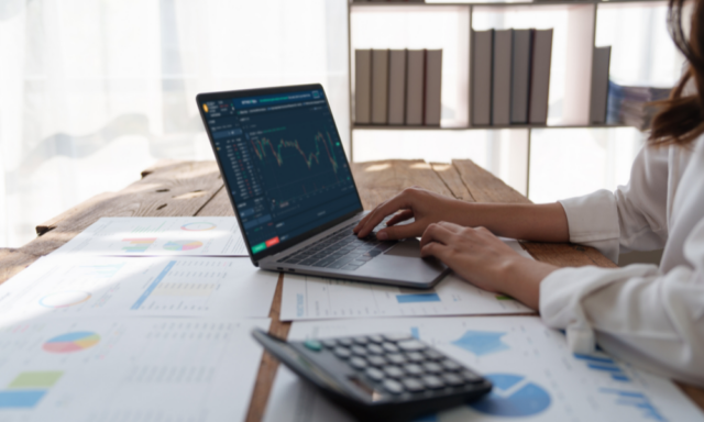 Woman working at a table with a laptop, calculator, and printed charts and graphs