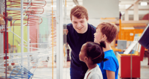 Why You Need More Teens in Your Museum Young adult talking to 2 kids in front of a science exhibit